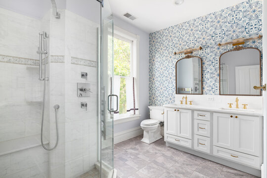 Fototapeta A bathroom with a blue wallpaper accent wall, gold mirror and faucets, grey cabinet, tiled flooring, and a walk-in shower with a chrome showerhead.