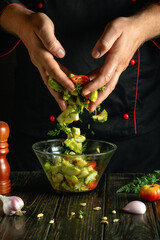 Experienced cook prepares salad in a bowl on the kitchen table with fresh vegetables. Mixing vegetable ingredients with hands.