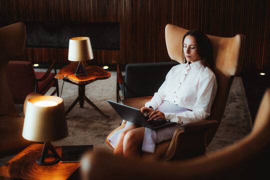 A caucasian woman in a formal white blouse and light purple skirt sits relaxed in an armchair with her eyes closed, using a laptop on her lap in a modern, stylish lounge setting
