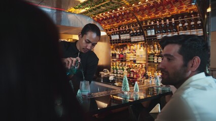 Bartender pouring mixed blue lagoon cocktail liqueur into cooling glass at fronted guest decorated pine light christmas theme, making special festive tasty in couple or friend at bar luxury. Vinosity.