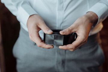 A man is holding a black box in his hand. The box is rectangular and has a silver color. The man is wearing a white shirt and gray pants. Concept of mystery and anticipation