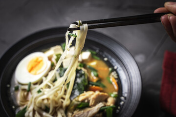 Chopsticks hold udon noodles over a plate of Miso Soup with tofu cheese, chicken, green onions and...