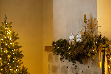 Stylish christmas decorated fireplace mantel with fir branch, bells, ribbon and garland light on background of christmas tree in modern living room. Atmospheric eve at home. Merry Christmas