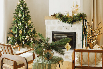 Stylish christmas decorated living room with christmas tree and modern fireplace mantel with fir branch, bells, ribbon and garland light. Atmospheric xmas holidays at home. Merry Christmas!
