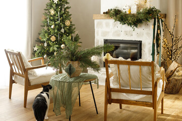 Cute cat sitting in stylish christmas decorated living room with christmas tree and modern fireplace mantel with fir branch, bells and garland light. Atmospheric holidays at home. Merry Christmas!