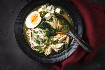 Japanese miso soup with udon noodles, tofu cheese, chicken, green onions and sesame seeds in a black bowl with a spoon and a burgundy napkin on a dark background. Top view.
