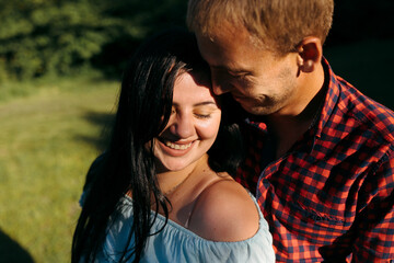 A man and a woman are hugging each other in a grassy field. The woman is smiling and the man is looking at her. Scene is happy and affectionate