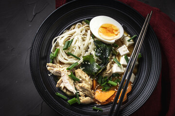 Japanese miso soup with udon noodles, tofu cheese, chicken, green onions and sesame seeds in a black bowl with chopsticks and a burgundy napkin on a dark background. Top view. Copyspace