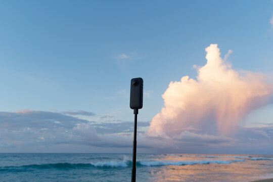 360 camera stands on tripod near ocean at sunset. 