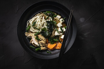 Japanese Miso Soup with udon noodles, tofu cheese, chicken, green onions and sesame seeds in a black bowl with chopsticks on a dark background. Top view. Copyspace