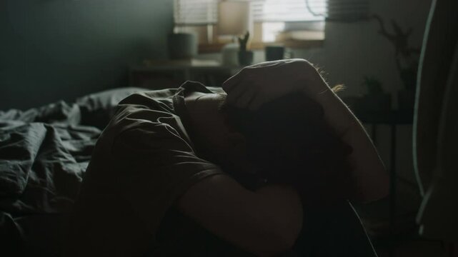 Sleepy young man putting head on knees, running hand through hair and rubbing cheek while sitting on floor by unmade bed in his room