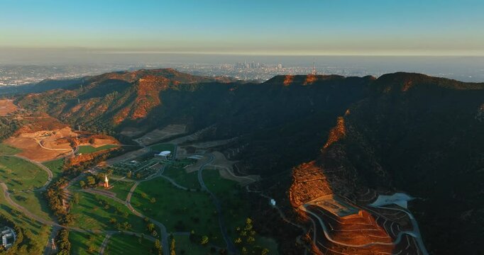 Griffith Park is shaded by the mountain at sunset. Drone rising over the scenery revealing the view on Los Angeles downtown at backdrop.