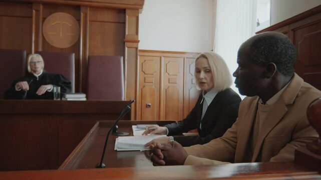 Caucasian female lawyer and her Black male client listening to judge, then she pointing at document and explaining it to him