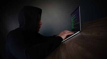 A hooded young guy types on a laptop displaying green code in a dimly lit room, sitting at a wooden table, representing themes of hacking, cybersecurity, or digital privacy in a secretive setting.