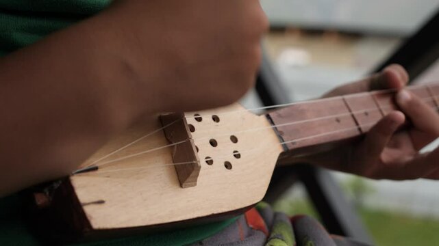 A young boy strums a traditional childrens size musical instrument. Closeup shot of the child hands joyfully playing a musical instrument with strings outdoors.
