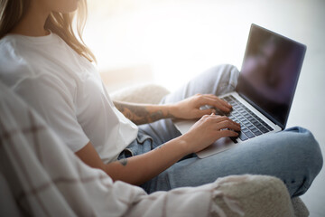Naklejka premium Close up image of woman hands while she sits comfortably in a cozy chair, working on her laptop in a bright, sunlit room. She appears focused and relaxed, embodying a modern, remote work lifestyle.