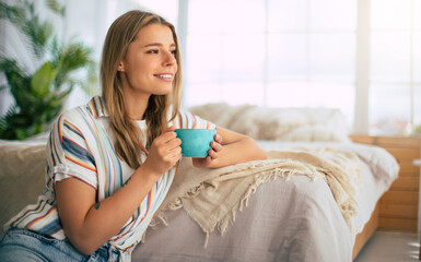 Cute woman smiles warmly while holding blue cup, sitting in a cozy, bright bedroom. She wears a casual striped shirt, with soft natural light and greenery in the background, creating a relaxed vibe