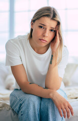A young woman sits on her bed, looking worried and holding her head. The bright room contrasts her expression of concern, suggesting stress, anxiety, or a health issue. Mental health or headache