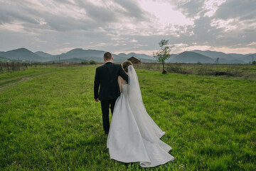 A bride and groom are walking through a field on their wedding day. The sky is cloudy and the couple is surrounded by a beautiful landscape