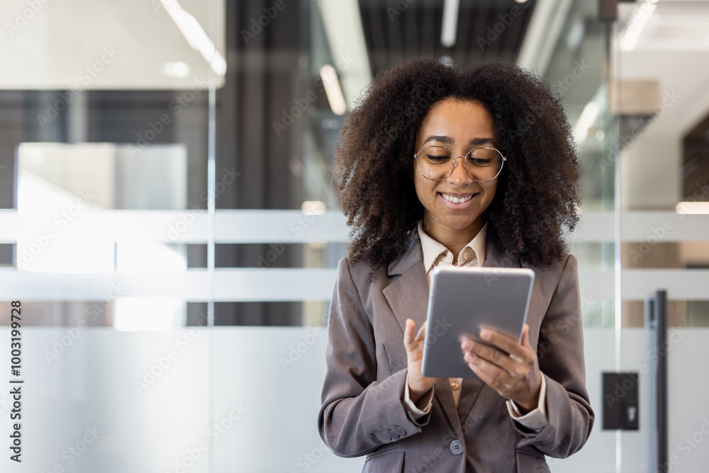 Wall mural smiling young african american businesswoman standing in suit in office and using tablet