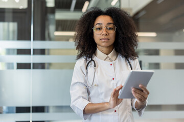 Portrait of serious young African American female doctor in uniform and glasses standing inside clinic, holding tablet and looking at camera