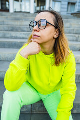 A young woman wearing a bright neon hoodie and pants sits on stone steps, deep in thought,...