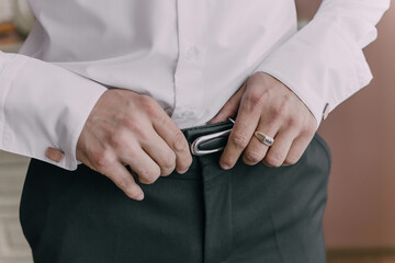 A man is adjusting his belt while wearing a white shirt and a gold ring. Concept of formality and attention to detail, as the man takes care to ensure his outfit is properly fitted