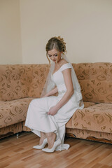 A bride is sitting on a couch, putting on her wedding shoes. The couch is brown and has a pattern on it. The bride is wearing a white dress and has her hair in a bun. She is focused on her shoes