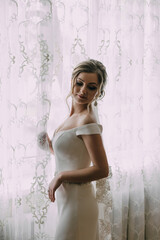 A woman in a wedding dress stands in front of a curtain, looking out the window. The scene is serene and peaceful, with the woman's elegant attire and the soft light coming through the window