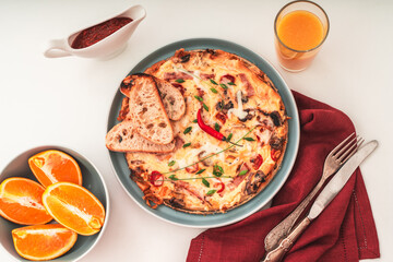 Italian frittata or omelette with tomatoes, cheese and toast on a blue plate with cutlery and a napkin on a white table and a glass of orange juice. Healthy breakfast concept. Selected Focus