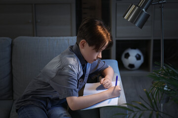 Little boy sitting on sofa and doing homework at home in evening