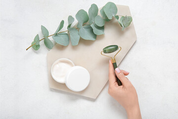 Female hand with stone face massage roller and cosmetic cream on light background