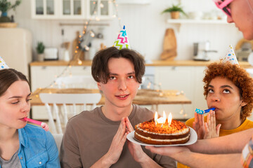 Make a wish. Man wearing party cap blowing out burning candles on birthday cake. Happy Birthday party. Group of friends wishes guy happy birthday. People celebrating birthday with party at home