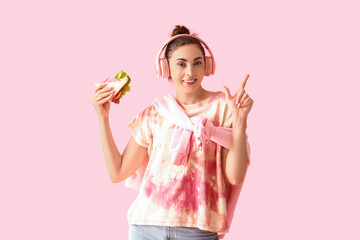 Young woman in headphones with tasty sandwich pointing at something on pink background