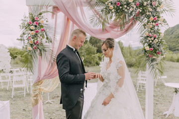 A bride and groom are getting married under a pink arch. The bride is wearing a white dress and the...