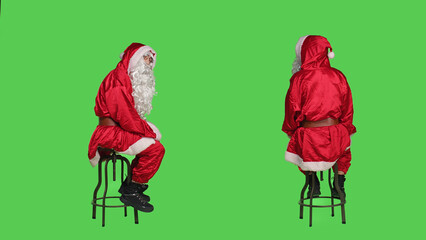 Santa claus on chair looking around, posing over greenscreen backdrop during christmas holiday. Model portraying saint nick character for december traditional celebration event.
