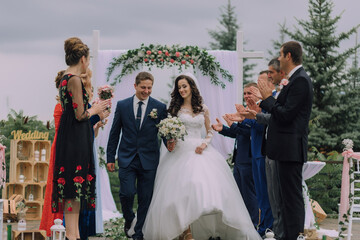 A bride and groom are walking down the aisle of a wedding ceremony. The bride is wearing a white...