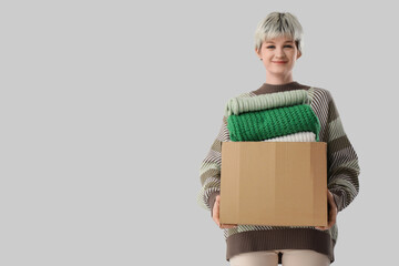 Beautiful young woman holding cardboard box with stacks of clothes on grey background