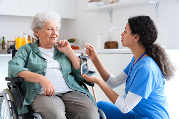 Young African-American female medical worker giving hearing aid elderly woman on wheelchair in...