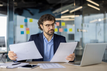 Mature financier examining paperwork in modern office environment with natural lighting and glass walls, emphasizing professionalism.