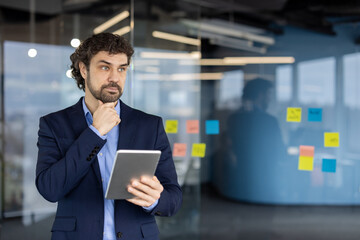 Thoughtful businessman holding tablet while standing in modern office with sticky notes on glass wall