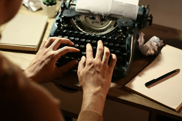 Young writer with vintage typewriter and lamp typing at table in room, closeup