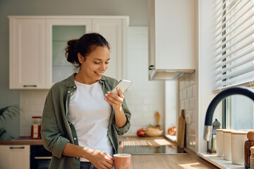 Happy Hispanic woman using cell phone while drinking coffee in kitchen.