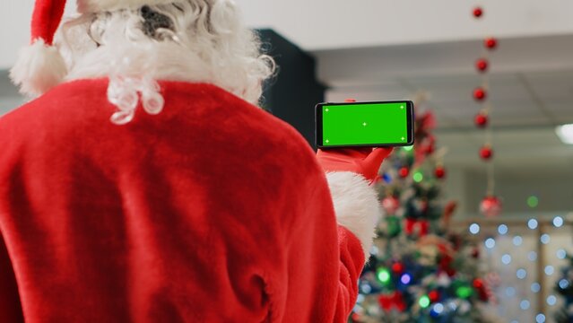 Employee in festive ornate shopping mall wearing Santa Claus suit holding green screen phone, taking a break from job shift and watching videos on chroma key screen, close up