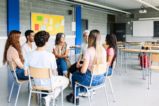 Young group of teenage students discussing in classroom sitting in circle with female teacher, giving an opinion on a debate topic. Education concept