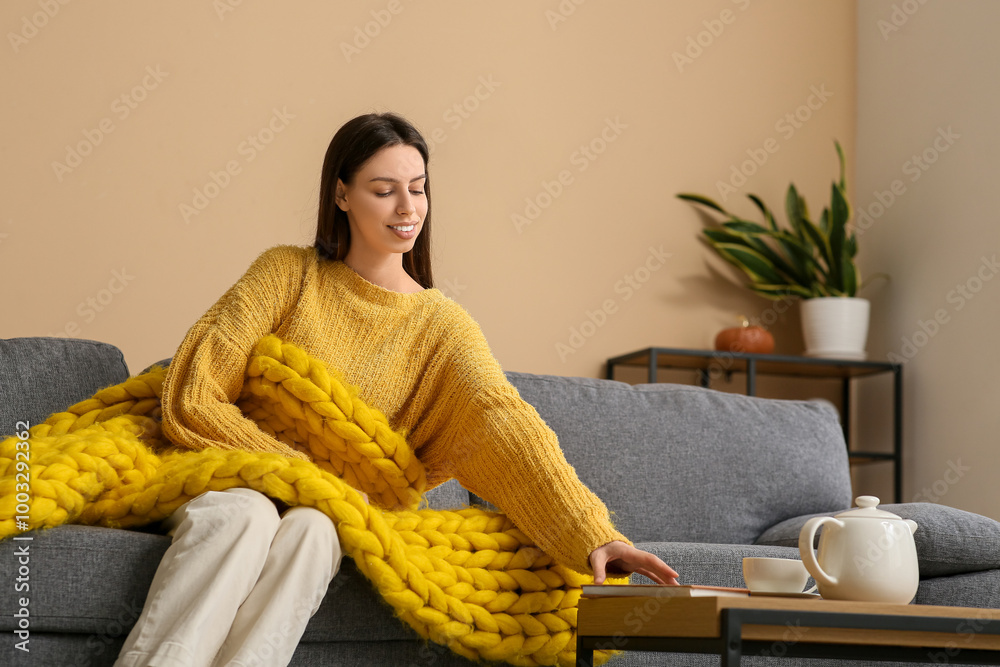 Poster Beautiful young woman with warm yellow plaid sitting on sofa and taking book from table in living room