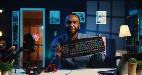 Influencer in dimly lit living room uses camera to film keyboard, mouse and headphones review for online streaming platforms. Online star hosts internet show, unboxing wired gaming peripherals