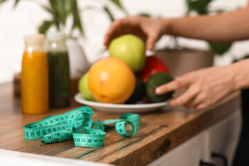 Tape measure on counter against woman with healthy food in kitchen, closeup. Diet concept
