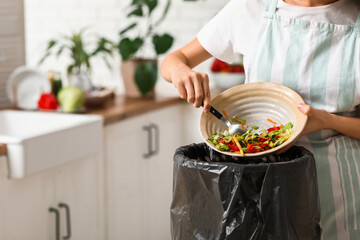 Young woman throwing organic food into trash bin in kitchen, closeup. Waste recycling concept