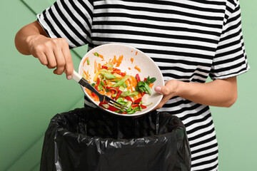 Young woman throwing organic food into trash bin on green background, closeup. Waste recycling concept